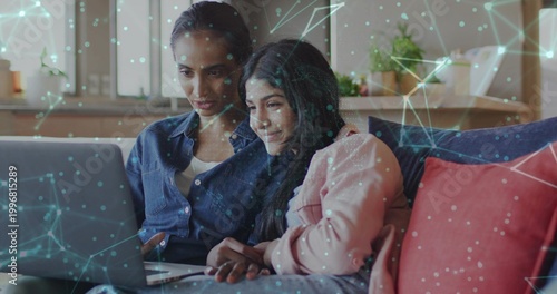 Sharing laptop, mother and daughter sitting on blue sofa, her in pink hoodie with tech overlay