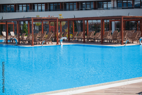 Wide angle view of luxury hotel resort building facade with large blue swimming pool and wooden pergolas. Summer vacation architecture background