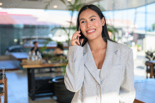A smiling Asian woman from Thai Isan talking on a smartphone in a professional cafe.
