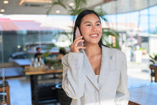 A smiling Asian woman from Thai Isan talking on a smartphone in a professional cafe.