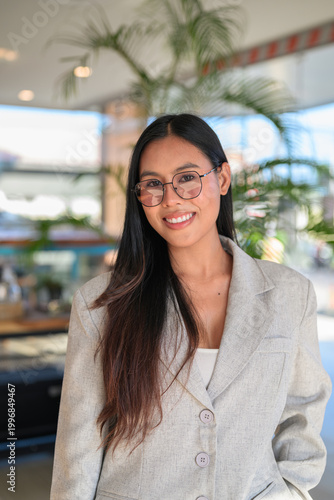 A smiling Asian woman from Thai Isan standing in a professional cafe wearing glasses.