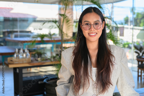 A smiling Asian woman from Thai Isan standing in a professional cafe wearing glasses.