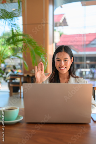 An authentic Asian woman from Thai Isan waving during a professional video call on a laptop in a cafe.