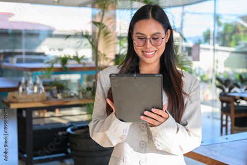A smiling Asian woman from Thai Isan using a tablet while wearing glasses in a professional cafe.