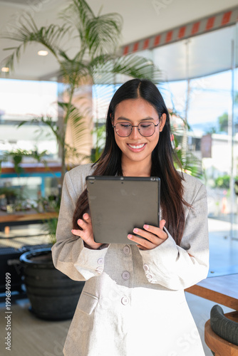 A smiling Asian woman from Thai Isan using a tablet while wearing glasses in a professional cafe