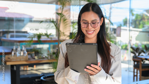A smiling Asian woman from Thai Isan using a tablet while wearing glasses in a professional cafe.