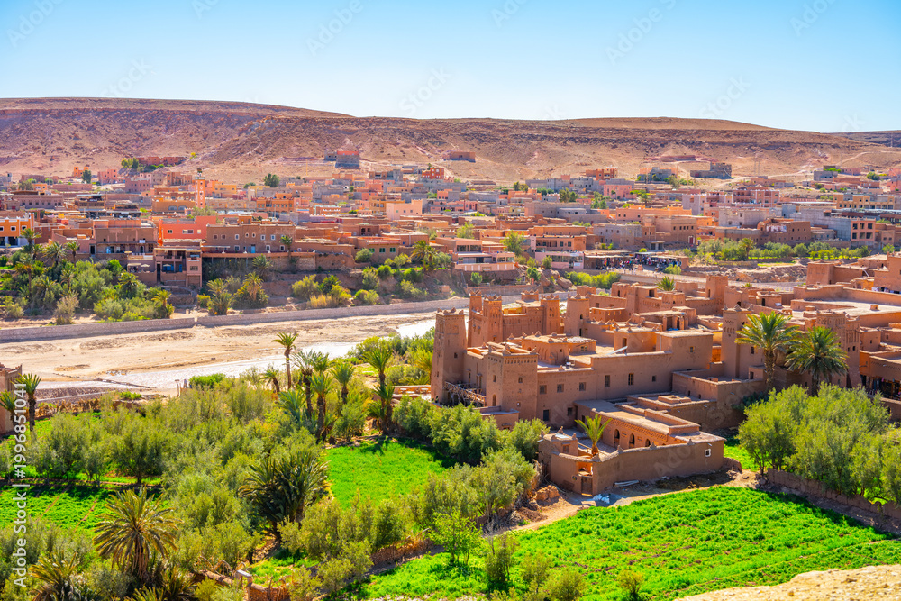 Obraz premium Ait Benhaddou stands along the caravan route. This site shows earthen architecture and traditional village life. Surrounding palm trees and green fields highlight its desert setting.