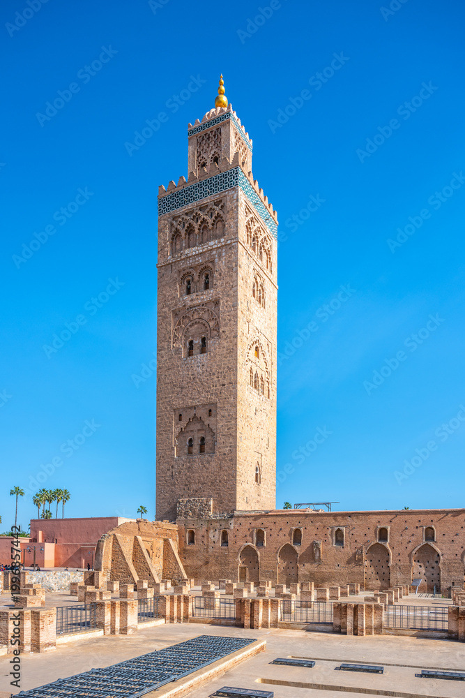 Obraz premium Koutoubia Mosque is located in Marrakesh, Morocco. The tall tower reaches up to the blue sky. Visitors admire the mosque's architecture during a sunny day in the city.