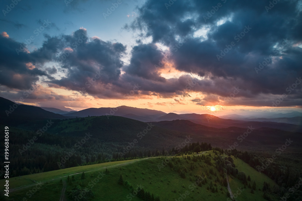 Obraz premium Aerial view breathtaking sunset over forest. Sun's rays break through dramatic clouds, casting golden glow across rolling hills and valleys. Distant mountains creating serene and picturesque scene.