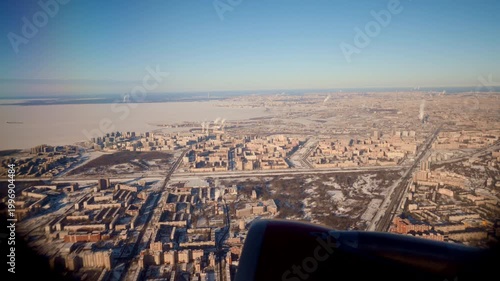 Aerial Airplane Window View Over City, Frozen Lake And Snowcovered Blocks Unfolding Below, Jet Engine In Foreground, Clear Blue Sky, Golden Morning Light, Lowaltitude Descent Toward Urban Coastline,