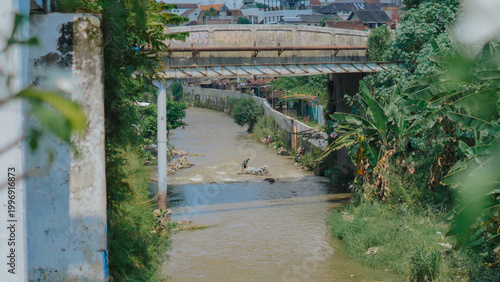 Konstfotografi A view of a murky river in a densely populated residential area, crossed by a large concrete bridge with pipes running alongside it