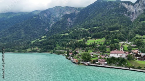 Drone flies forward over turquoise Lake Brienz revealing charming Swiss alpine village of Brienz with church, chalets, and dramatic misty mountains.