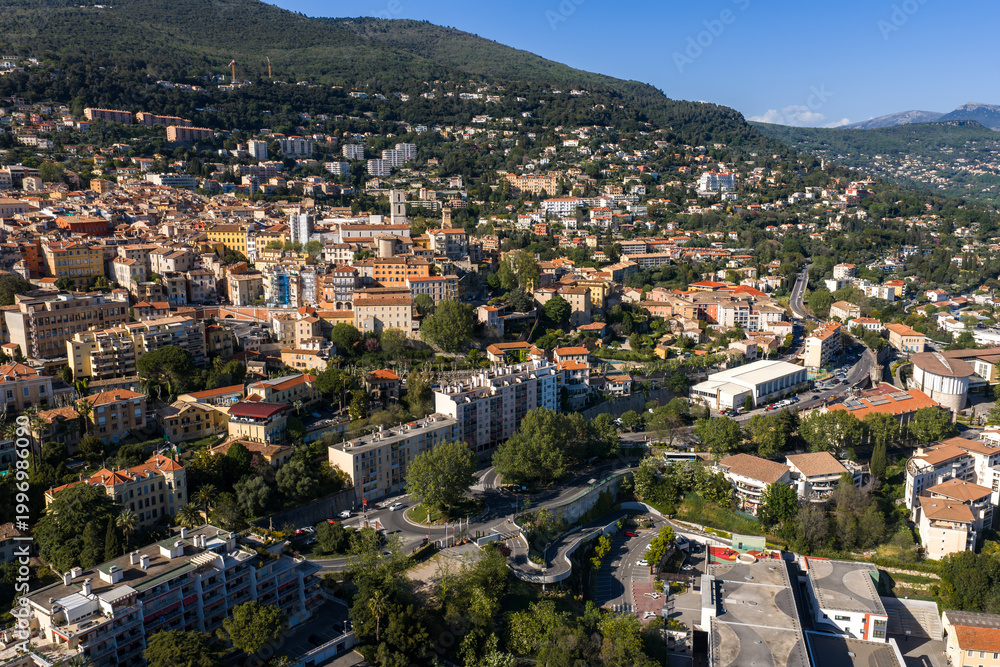 Fototapeta premium Aerial view of Grasse, France shows terracotta roofs on slopes, church towers, a roundabout, cypress and plane trees, and Alpine foothills in clear morning light.