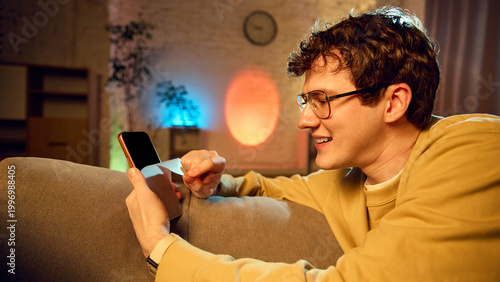 Fotografie Man peeling protective layer from new smartphone while sitting on sofa in cozy interior