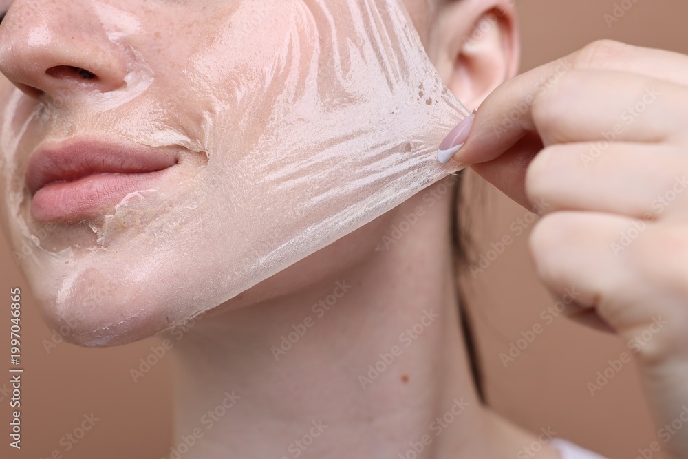 Fototapeta premium Beautiful woman peeling off facial mask on brown background, closeup