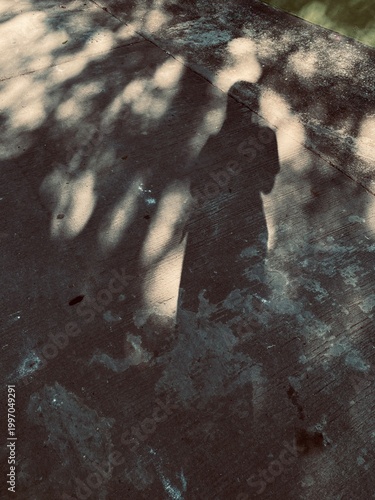 Concept of solitude and introspection with a person's shadow cast on a weathered pavement, mixed with light and shadow of tree leaves.