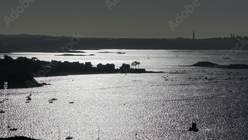Aerial view of glimmering waters reflecting the stark contrast of light and shadow upon the distant cityscape and rugged coastline, Carantec, Bretagne, France.