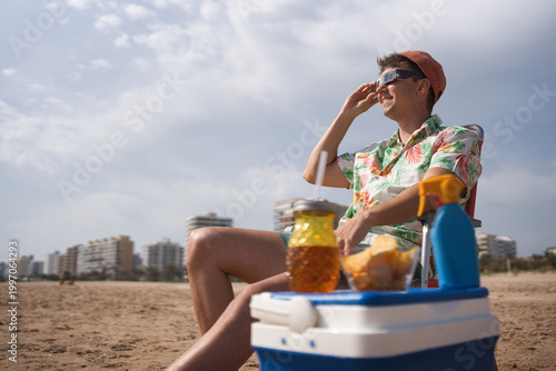 Man in hawaiian shirt and cap wearing protective eclipse glasses on an urban beach, sitting by a cooler with drink and snacks, enjoying a summer daytime solar eclipse