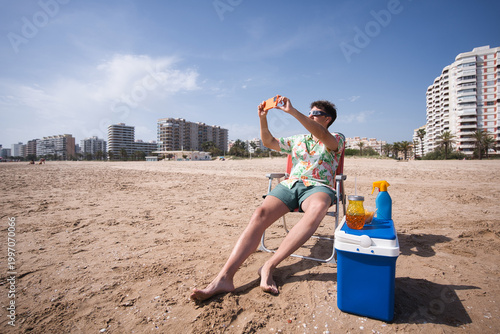 Man on beach chair in hawaiian shirt photographing a solar eclipse with smartphone, cooler and sunscreen beside him under clear summer sky, relaxing on vacation destination