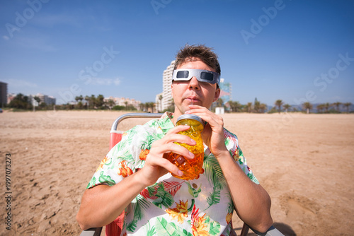 Bilde på lerret Man in hawaiian shirt wearing solar eclipse glasses sits in a beach chair, sippi
