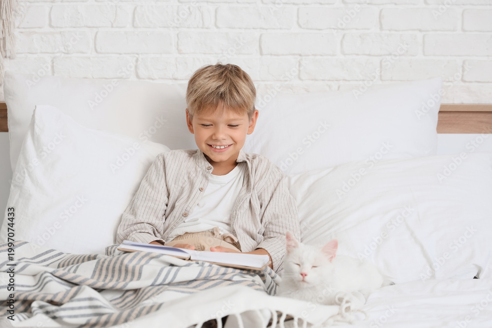Fototapeta premium Little boy with cat reading book under blanket in bedroom