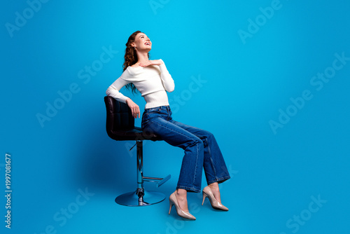 Happy young woman posing on a modern stool against a bright blue background in casual jeans and heels, stylish portrait conveying confidence, relaxation and contemporary fashion