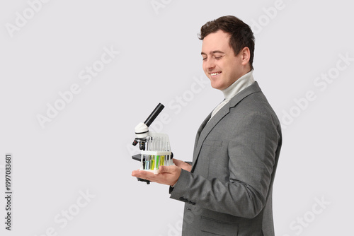 Young male teacher with microscope and chemical flasks on grey background