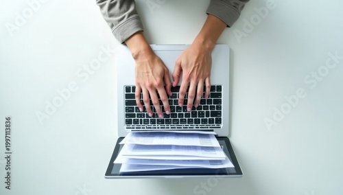 Vista superior de unas manos tecleando en un portátil con capas de documentos digitales que se expanden hacia afuera, escritorio blanco y limpio
