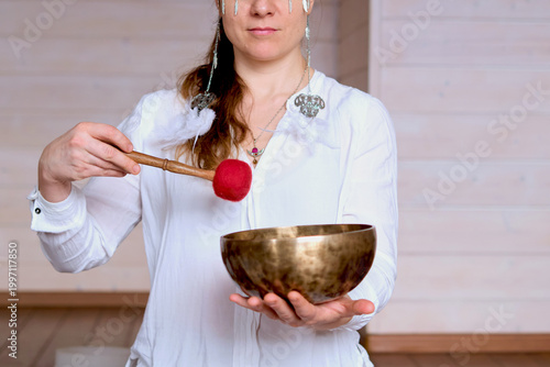 Close portrait of a calm woman with closed eyes holding a Tibetan singing bowl and red mallet in soft studio light. Sound meditation supports stress relief, deep rest, and emotional balance.