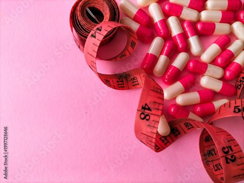 A pink measuring tape is coiled and spread across a light pink background. Nearby are numerous pink and white capsules, representing dietary supplements or weight loss pills.
