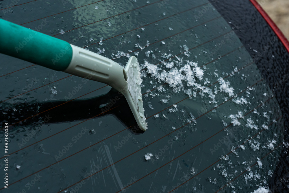 Obraz premium Removing Ice and Snow from a Car Rear Window with a Scraper Tool in Cold Weather Conditions