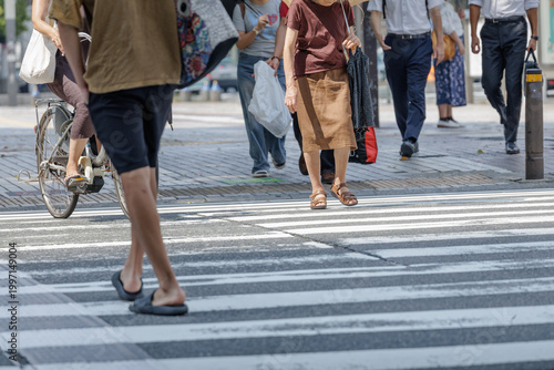 Konstfotografi Diverse crowd crossing sunlit street in Japan with cyclist gliding nonchalantly past showcases everyday commute, summer attire, movement and urban life with mixed-age shoppers and office workers