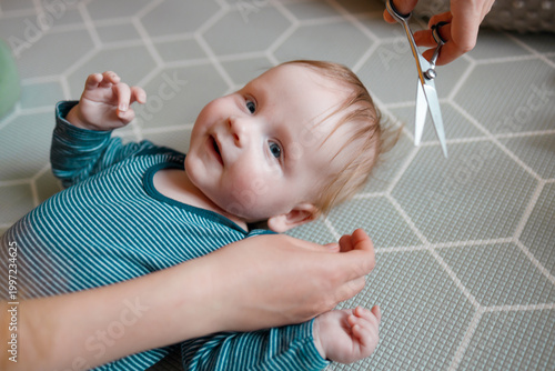 A cheerful infant boy getting his first haircut at home. The mother is carefully using professional scissors to trim the baby's hair while he lies on a play mat and smiles. Concept of first haircut