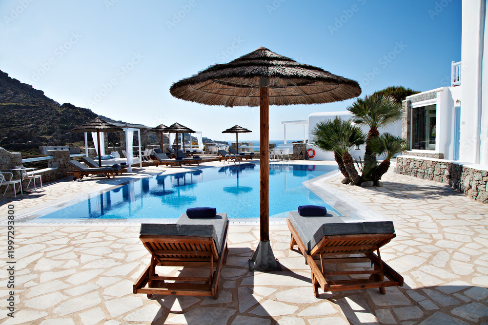 Obraz premium Luxury swimming pool with sunbeds and parasol in a Mediterranean resort setting. Bright summer scene with blue sky, clear water and relaxing atmosphere,.