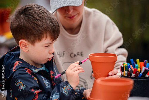 Little boy painting terracotta plant pot with pink marker while woman holds it outdoors