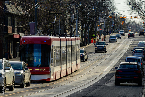 Modern red TTC streetcar moving through morning traffic on urban commercial street with overhead wires and spring trees.