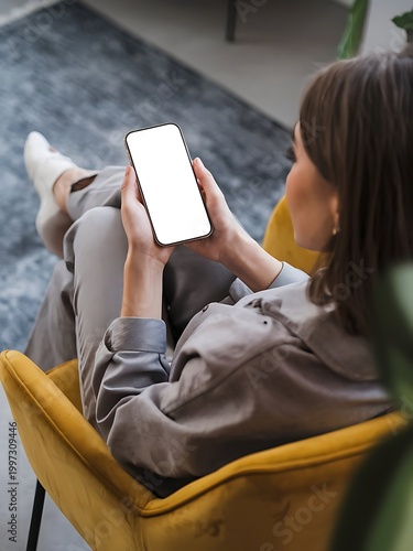 A woman holds phone with white screen sitting a yellow chair. Mockup