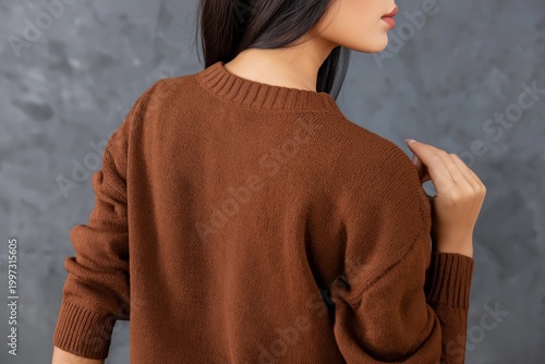 Woman in Brown Sweater Stands Against Gray Wall and Touches Shoulder With Hand