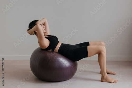 Slim caucasian woman with dark hair performing abdominal exercise on large fitness ball in modern pilates studio. Trendy sportswear and controlled movement highlight core strength balance and ifestyle