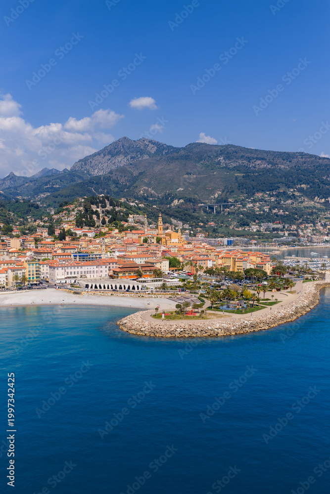 Fototapeta premium Aerial view of Menton, France shows pastel Old Town, Basilique Saint Michel bell tower, curved stone jetty, marina, Promenade du Soleil, turquoise sea, and Maritime Alps at midday.