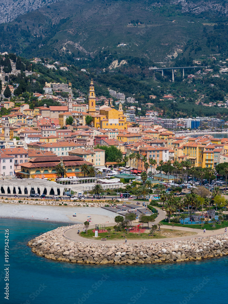 Fototapeta premium Aerial Menton, France, shows the Basilica of Saint Michel bell tower, terracotta roofs, marina and promenade, curved breakwater, sandy beach, and Alpine foothills in sun.