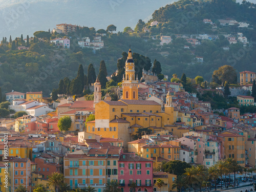 Bild auf Leinwand Sunlit Menton old town in France shows pastel buildings on a hillside, the twin towered Basilica of Saint Michel Archange, a yellow bell tower, and a seafront promenade