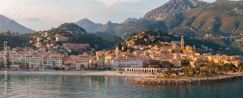 Aerial view of Menton, France, with the Basilica of Saint Michel, crescent promenade, curved breakwater, calm harbor, and hillside old town in late afternoon light.