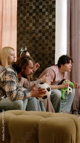 Friends joyfully react while watching a soccer game on a couch, holding soccer balls and showing excitement in a stylish living room