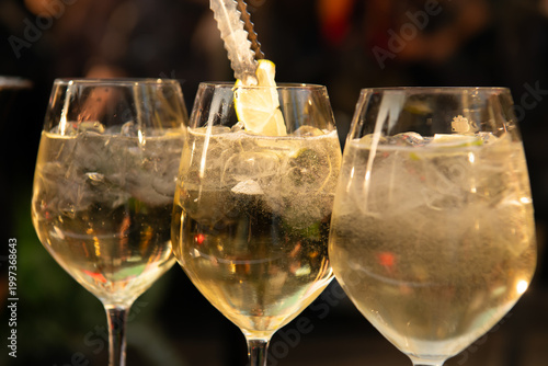 Close-up of three sparkling cocktails with ice and lemon garnish in elegant glasses, glowing in warm bar lighting, creating a festive and sophisticated evening atmosphere.