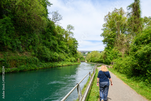 Woman walking along a peaceful riverside path near the Adda River, surrounded by lush greenery and calm turquoise water under a bright sky.