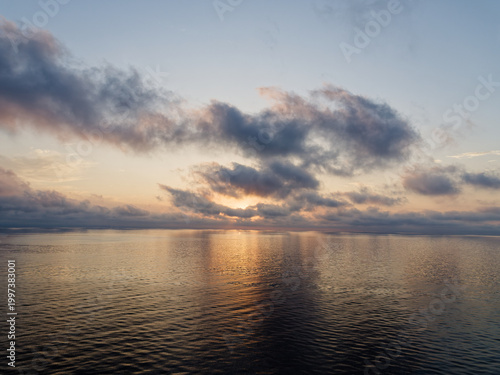 Dramatic sunset over a calm Mediterranean Sea, with golden sunlight reflecting on the water surface beneath a moody cloudscape of soft pink and blue-grey clouds, viewed from open sea.