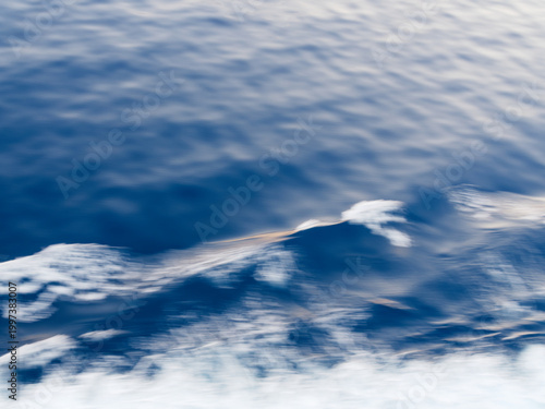 Abstract blue water surface texture with gentle ripples and soft sky reflections on the calm Mediterranean Sea, motion blurred soft white surf at the bottom