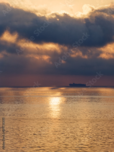 Dramatic sunrise over a calm Mediterranean Sea, with golden sunlight reflecting on the water surface beneath a moody cloudscape, in the distance the silhouette of a panamax container ship is visible