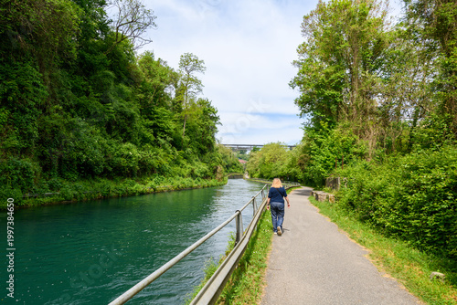 Woman walking along a peaceful riverside path near the Adda River, surrounded by lush greenery and calm turquoise water under a bright sky.